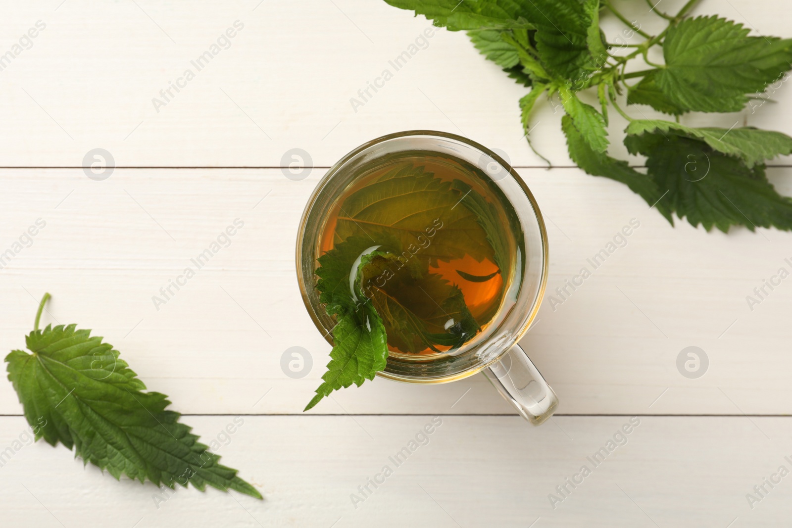 Photo of Glass cup of aromatic nettle tea and green leaves on white wooden table, flat lay