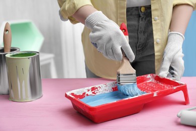 Photo of Woman taking light blue paint with brush from tray at pink wooden table indoors, closeup