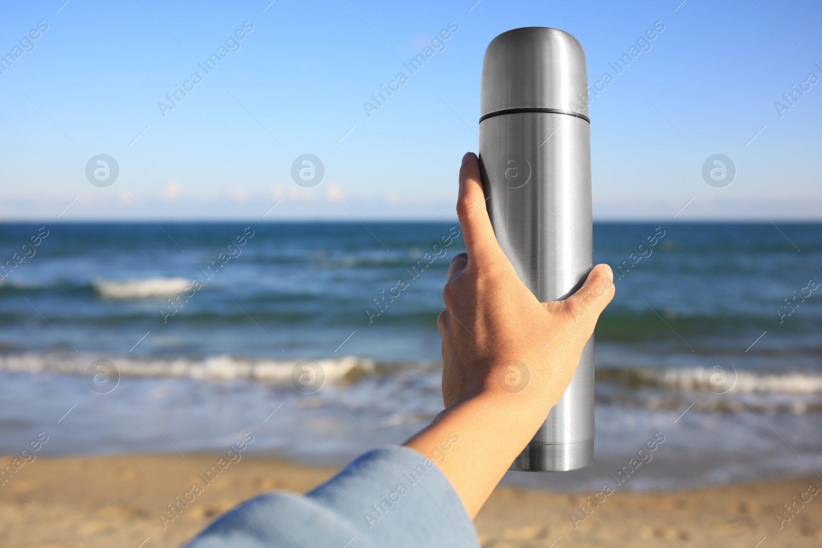 Photo of Woman holding metallic thermos with hot drink on beach near sea, closeup. Space for text