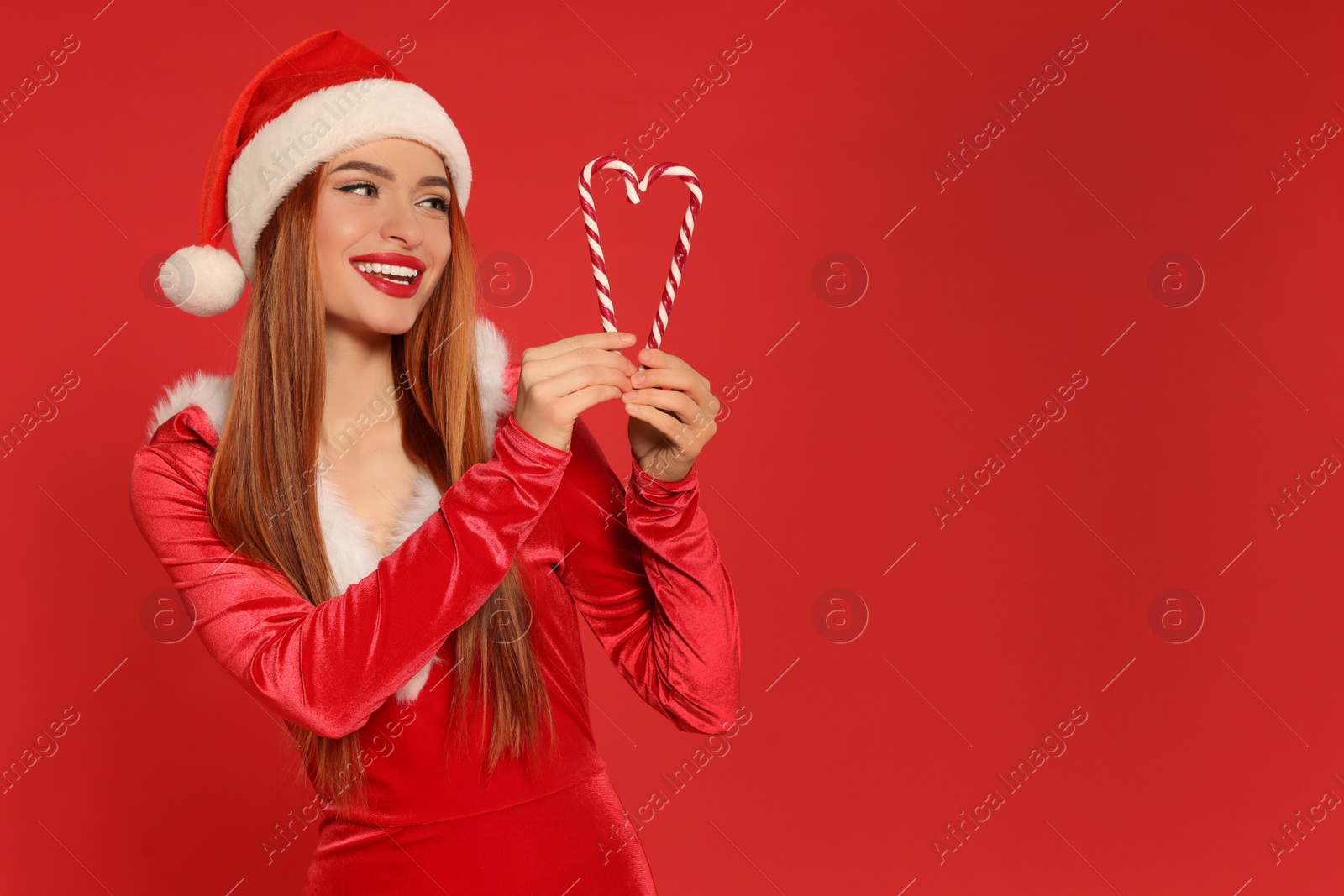 Photo of Young woman in Santa hat with candy canes on red background, space for text. Christmas celebration