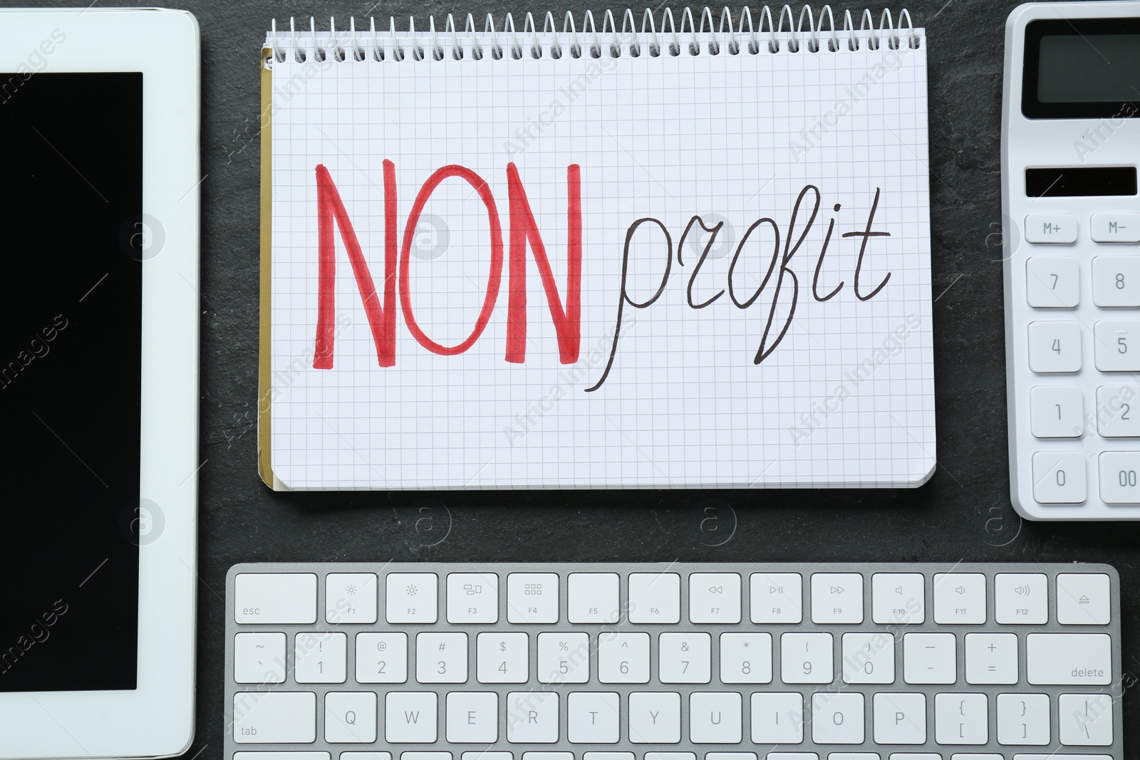 Photo of Notebook with phrase Non Profit, calculator, computer keyboard and tablet on black table. flat lay