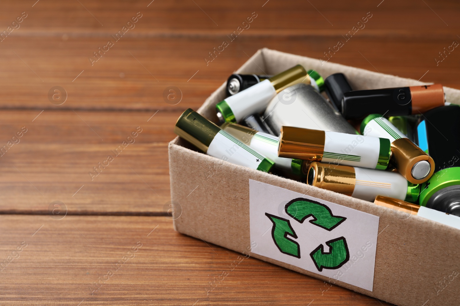 Image of Used batteries in cardboard box with recycling symbol on wooden table, closeup. Space for text