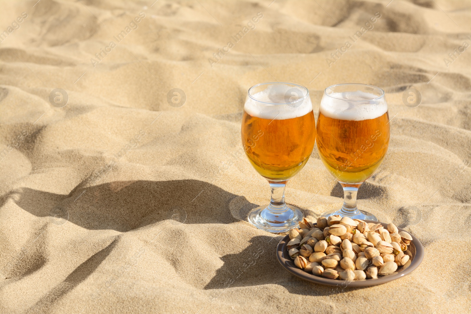 Photo of Glasses of cold beer and pistachios on sandy beach, space for text