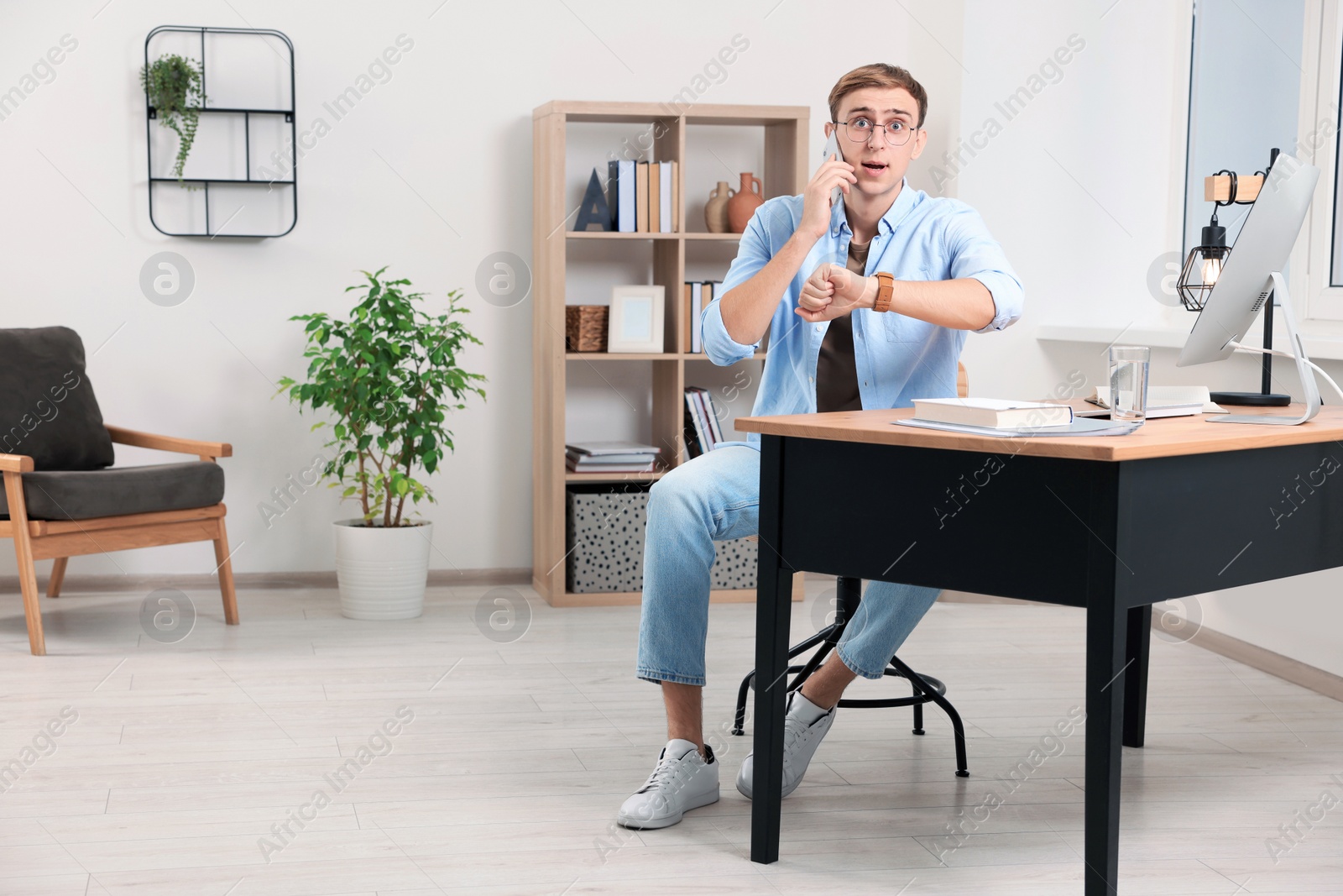 Photo of Emotional young man checking time while talking on phone in office. Being late