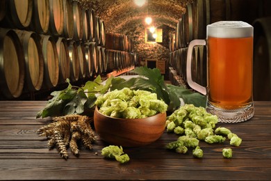 Glass of tasty light beer, fresh hops and wheat spikes on wooden table in cellar