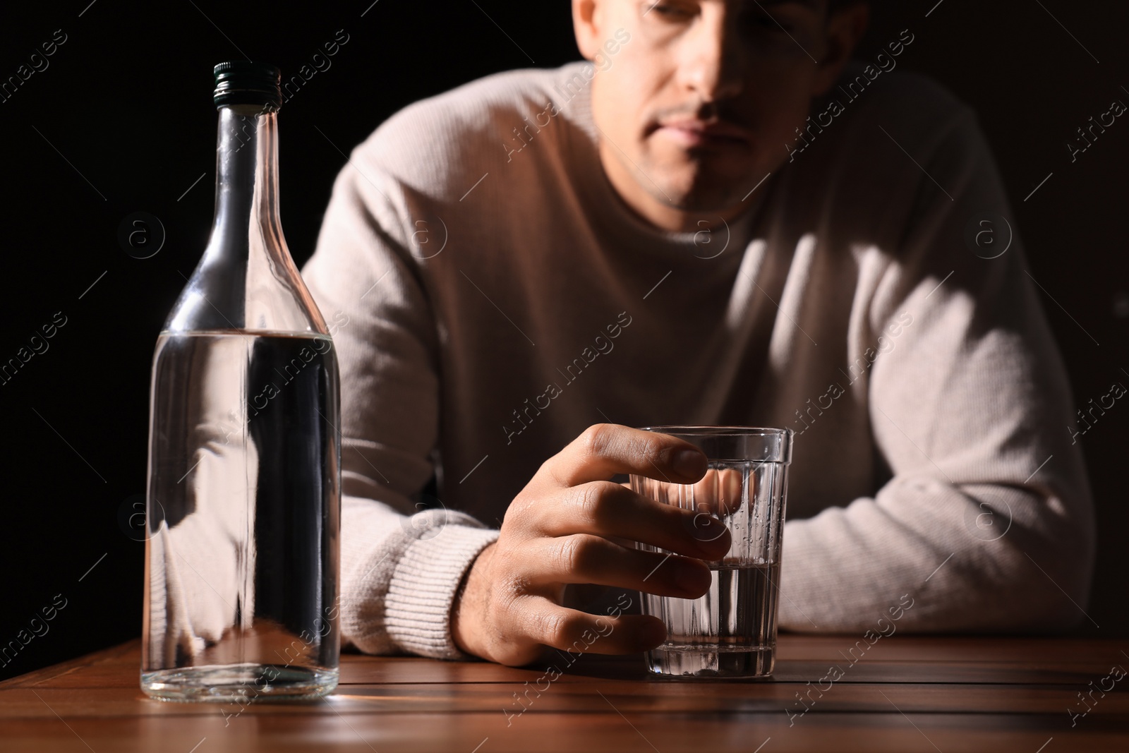 Photo of Addicted man with alcoholic drink at wooden table against black background, closeup