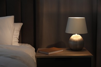 Stylish lamp and book on bedside table near bed indoors