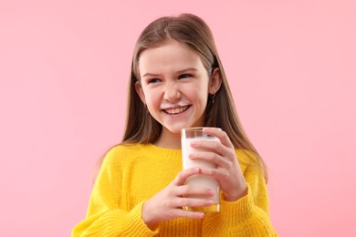 Happy little girl with milk mustache holding glass of tasty dairy drink on pink background