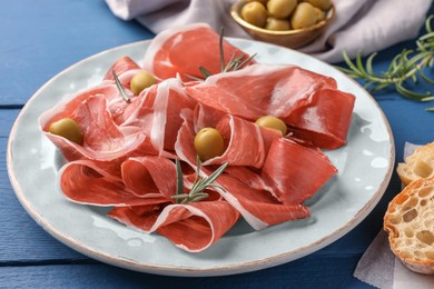 Photo of Slices of tasty cured ham, rosemary and olives on blue wooden table, closeup