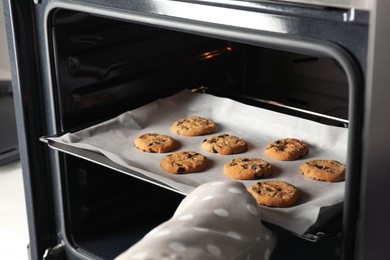 Photo of Woman taking out delicious chocolate chip cookies from oven, closeup