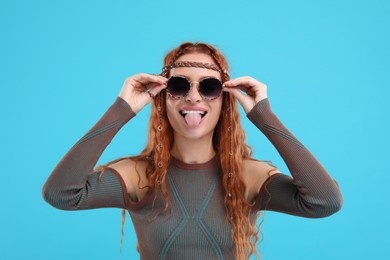 Photo of Stylish young hippie woman in sunglasses showing her tongue on light blue background