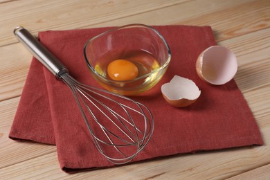 Photo of Metal whisk, raw eggs in bowl and shells on wooden table, closeup