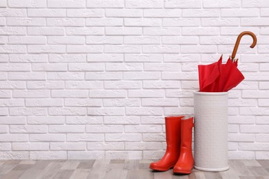Photo of Red umbrella in holder and rubber boots near white brick wall indoors. Space for text
