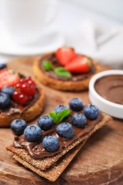 Photo of Fresh rye crispbreads with chocolate spread and blueberries on wooden board, closeup
