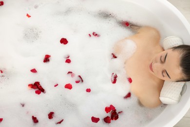 Photo of Woman taking bath in tub with foam and rose petals, top view