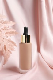 Photo of Bottle of skin foundation and decorative plant on pink cloth. Makeup product