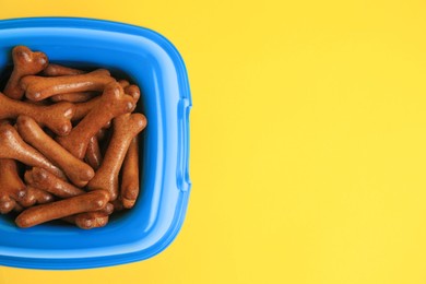 Photo of Blue bowl with bone shaped dog cookies on yellow background, top view. Space for text