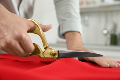 Photo of Professional tailor cutting red fabric with scissors in workshop, closeup