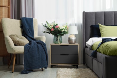 Photo of Large comfortable bed, armchair and beautiful bouquet on bedside table in room. Bedroom interior