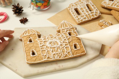 Photo of Woman decorating gingerbread house part with icing at white table, closeup