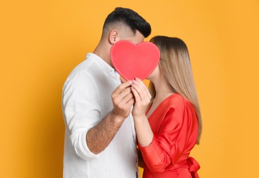 Photo of Lovely couple kissing behind decorative heart on yellow background. Valentine's day celebration