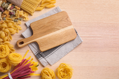 Photo of Different types of pasta and cutting board on wooden table, flat lay
