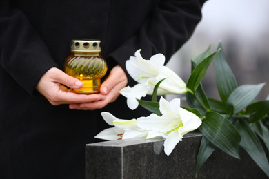 Photo of Woman with candle and white lilies near black granite tombstone outdoors, closeup. Funeral ceremony
