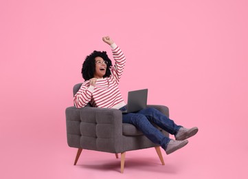 Emotional young woman with laptop sitting in armchair against pink background