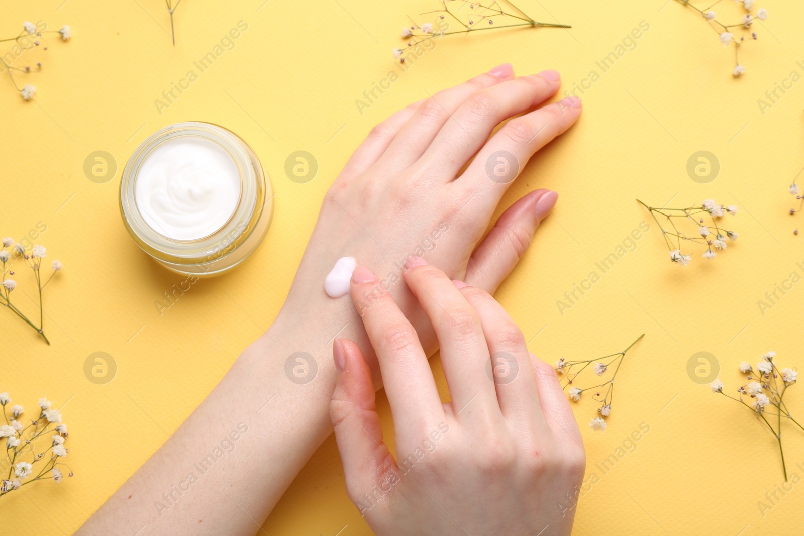 Photo of Woman applying hand cream and flowers on yellow background, top view