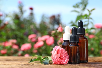 Photo of Bottles of essential oil and fresh rose on wooden table against blurred background. Space for text