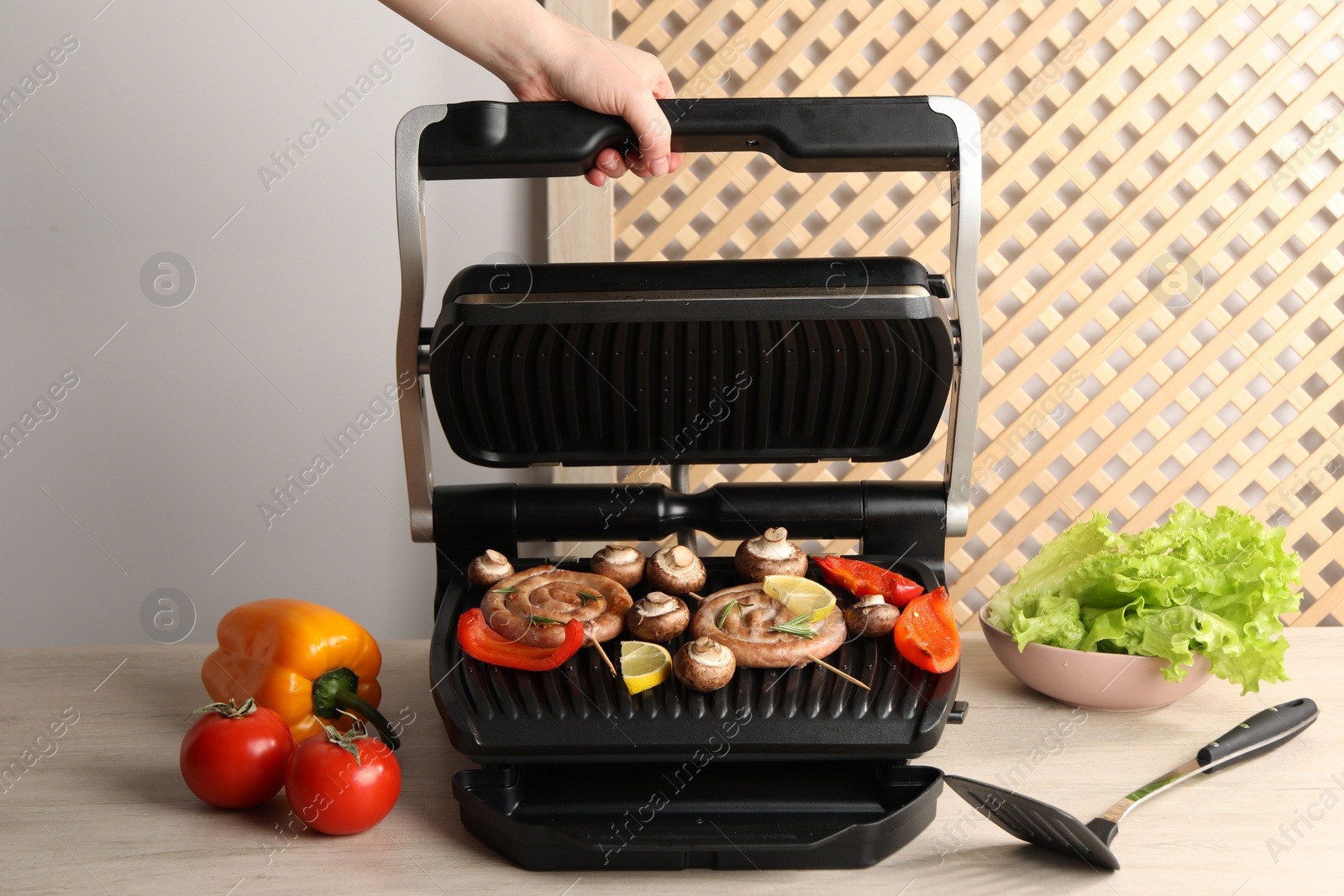 Photo of Woman cooking homemade sausages with bell peppers and mushrooms on electric grill at wooden table, closeup