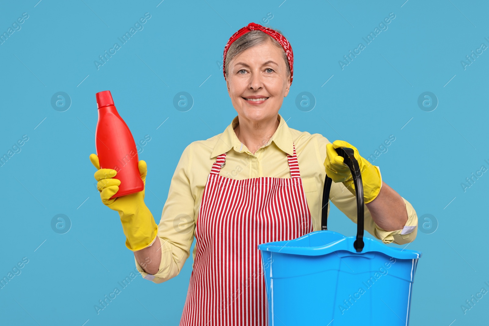 Photo of Happy housewife with bottle of detergent and bucket on light blue background