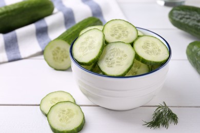 Photo of Cut cucumber in bowl, fresh vegetables and dill on white wooden table, closeup