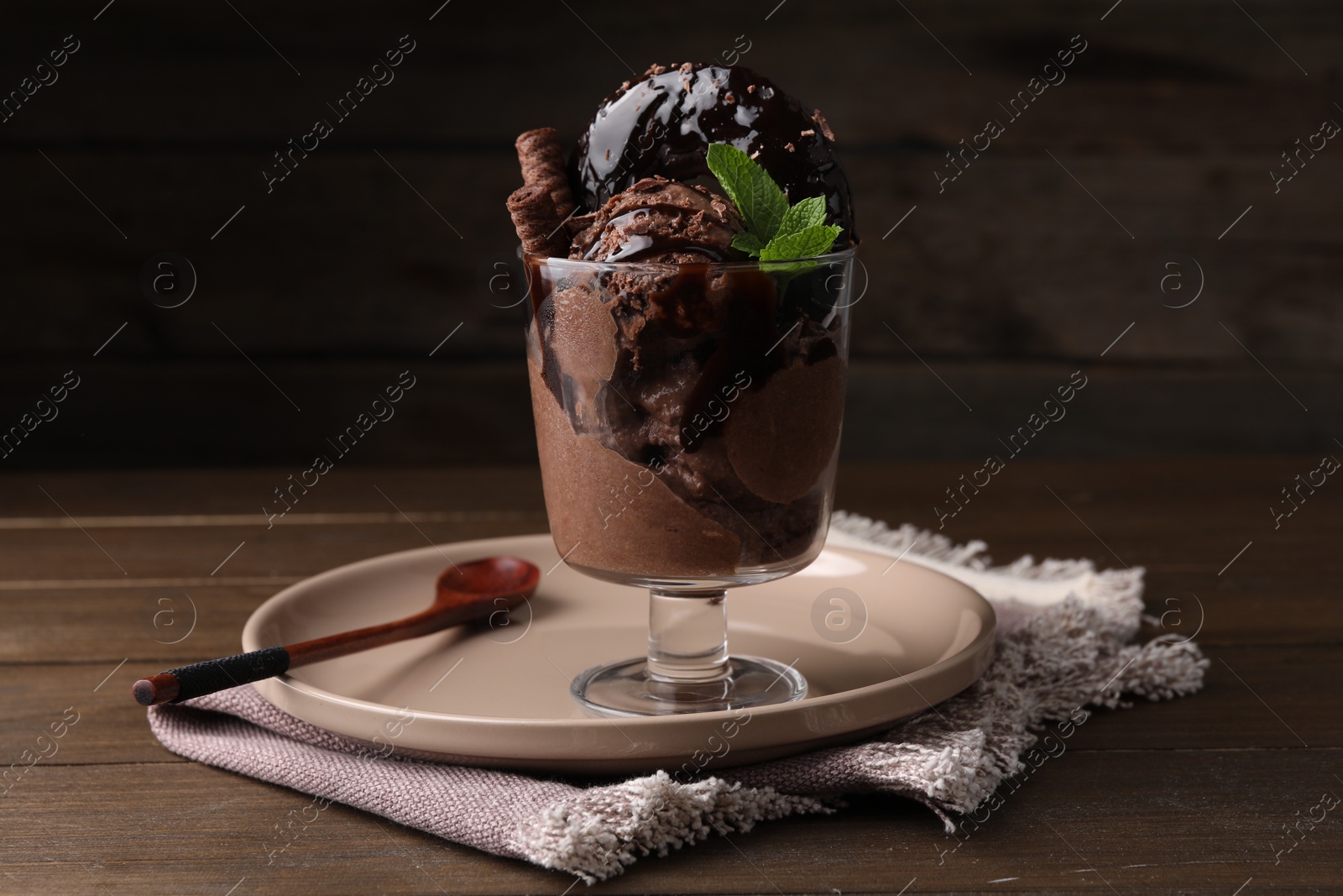 Photo of Delicious chocolate ice cream with wafer sticks, donut and mint in glass dessert bowl on wooden table