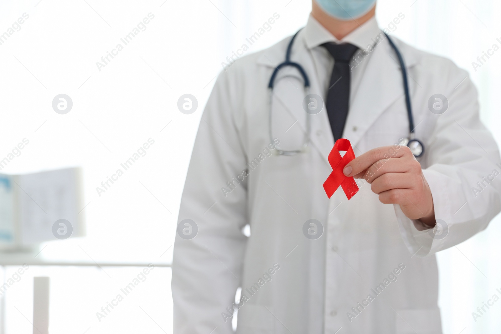 Photo of Doctor holding red ribbon on blurred background, closeup with space for text. AIDS awareness month