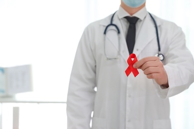 Photo of Doctor holding red ribbon on blurred background, closeup with space for text. AIDS awareness month