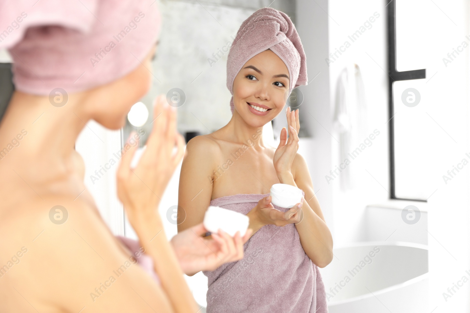 Photo of Young woman with towels applying facial cream near mirror in bathroom