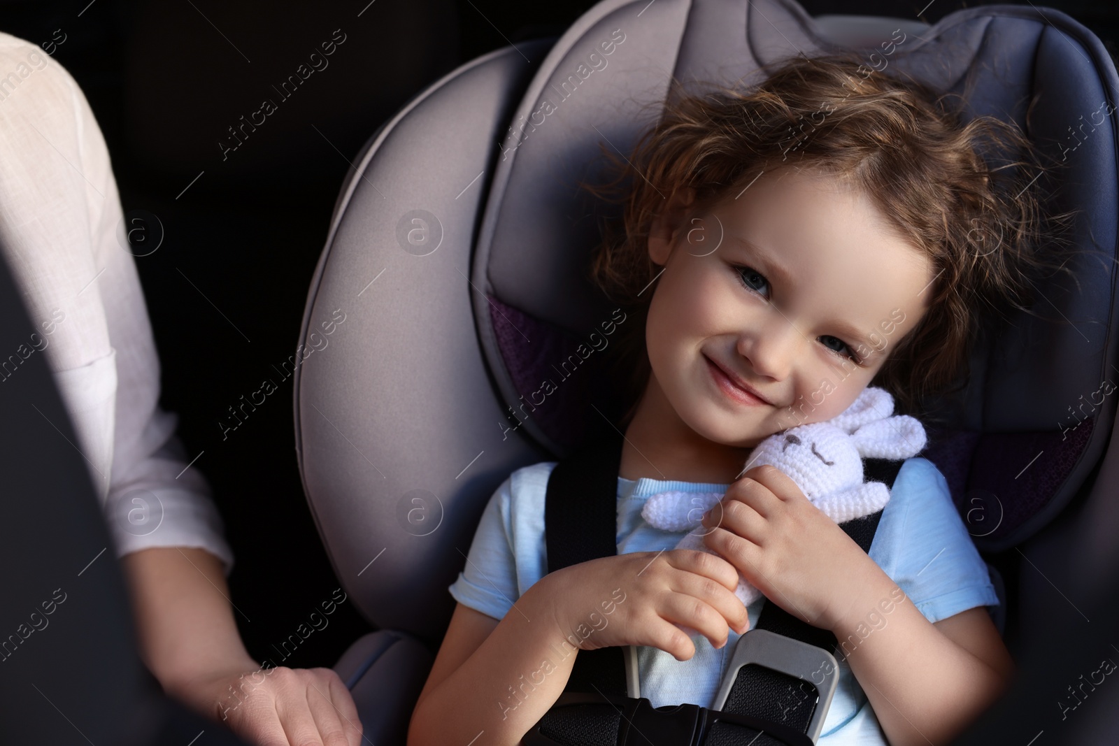Photo of Cute little girl with toy rabbit sitting in child safety seat near mother inside car