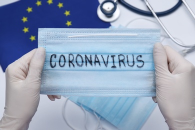 Photo of Doctor holding mask with word Coronavirus above medical items and European Union flag, closeup
