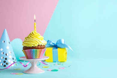 Photo of Delicious birthday cupcake with candle near gift box and party hat on color background, space for text