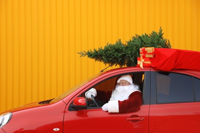Photo of Authentic Santa Claus with fir tree and bag full of presents on roof driving car against yellow background