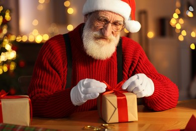 Photo of Santa Claus tying bow on gift box at his workplace in room decorated for Christmas