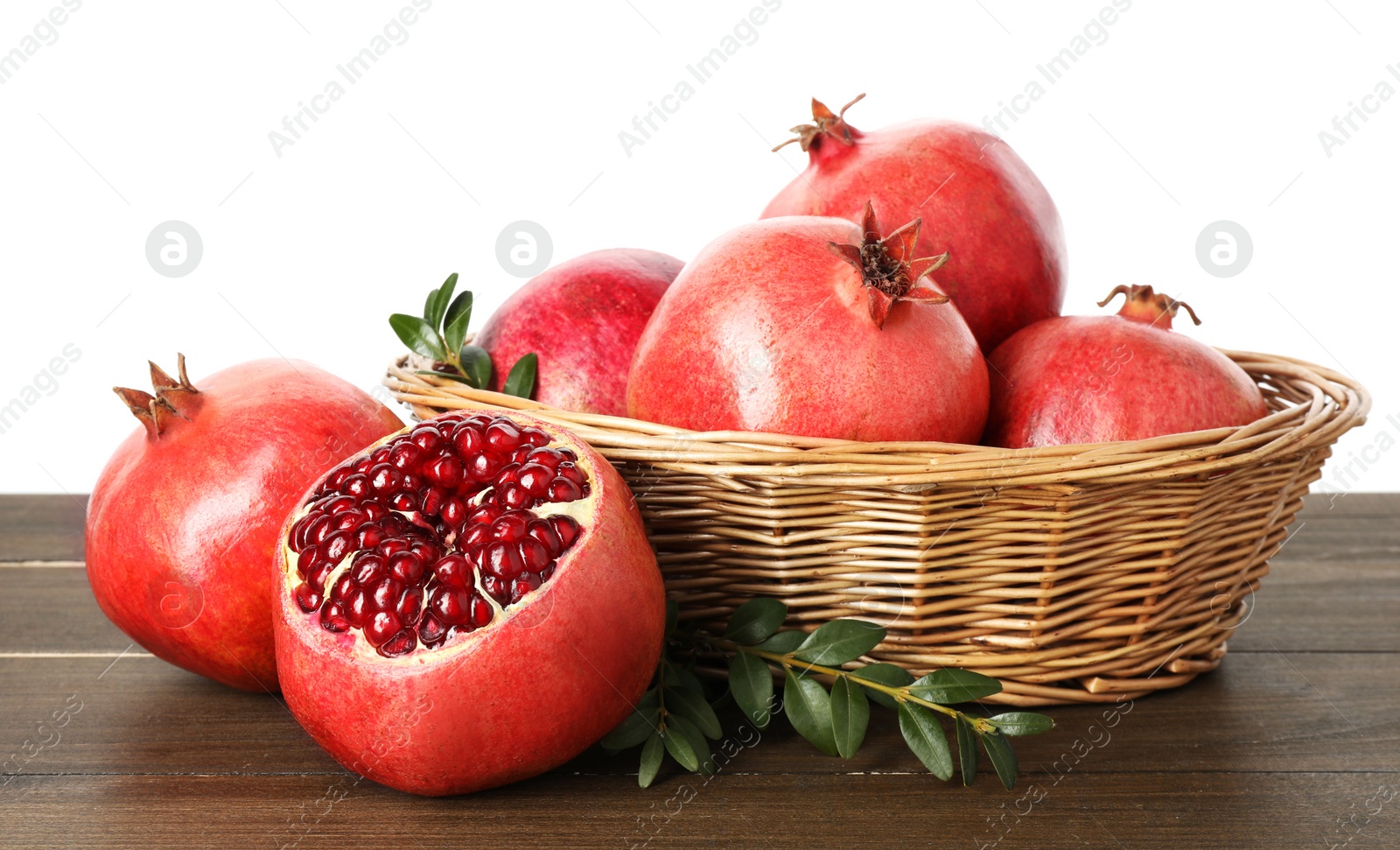 Photo of Fresh pomegranates in wicker basket and green leaves on wooden table against white background