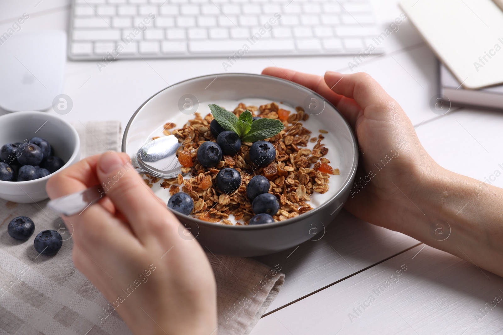 Photo of Woman holding bowl of tasty granola with blueberries at white wooden table, closeup