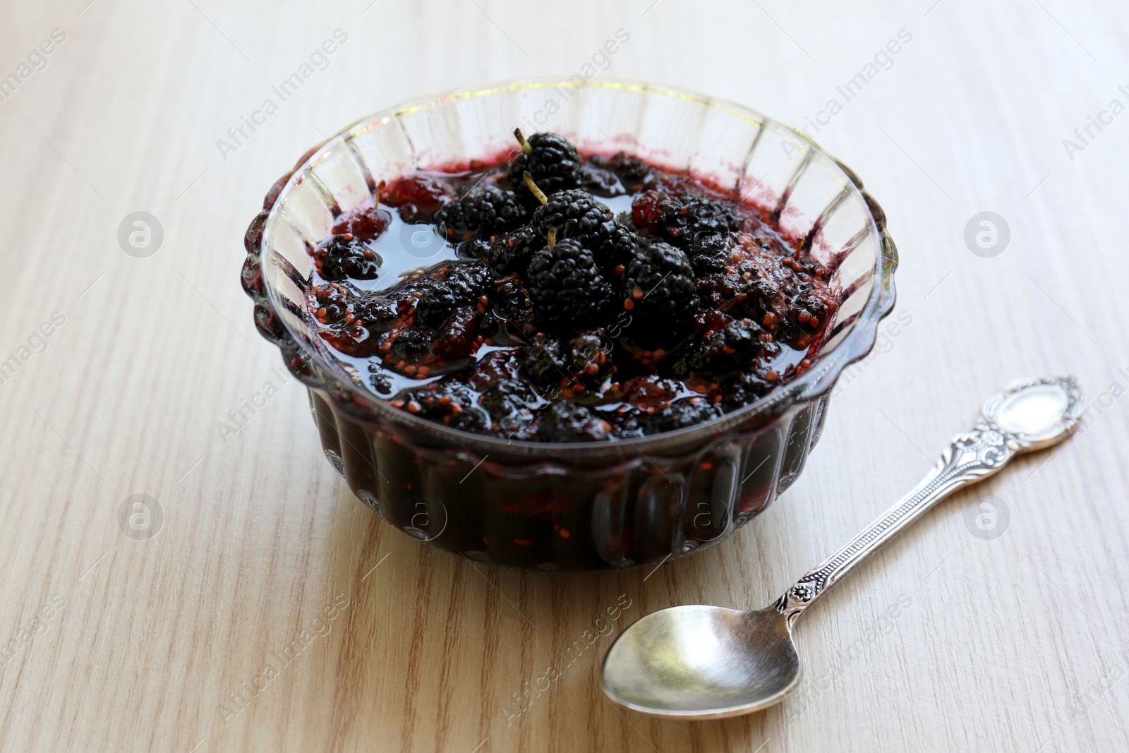 Photo of Bowl of sweet black mulberry jam and spoon on wooden table