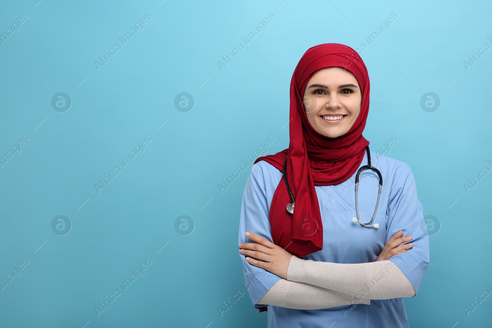 Photo of Muslim woman wearing hijab and medical uniform with stethoscope on light blue background, space for text