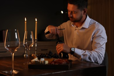 Photo of Romantic dinner. Man opening wine bottle with corkscrew at table indoors
