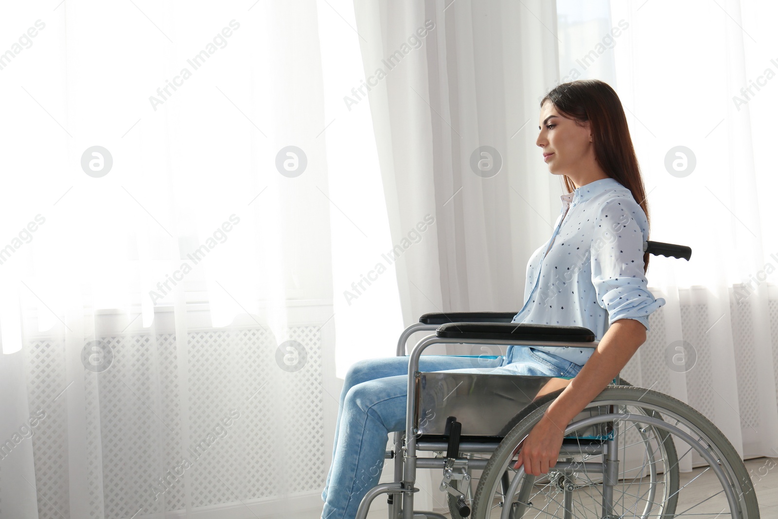 Photo of Young woman sitting in wheelchair near window indoors. Space for text