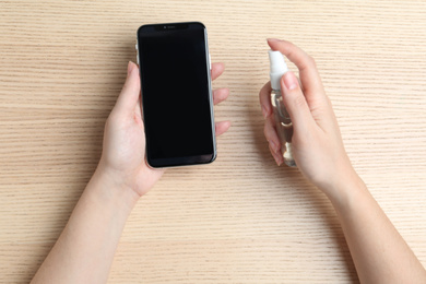 Photo of Woman spraying antiseptic onto smartphone at wooden table, top view