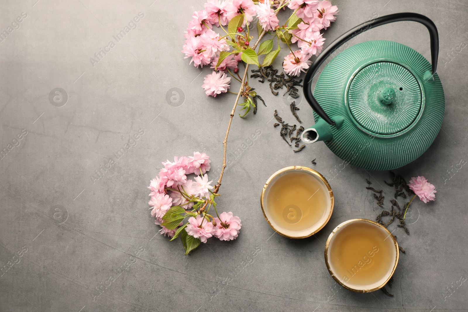 Photo of Traditional ceremony. Cups of brewed tea, teapot and sakura flowers on grey table, flat lay with space for text